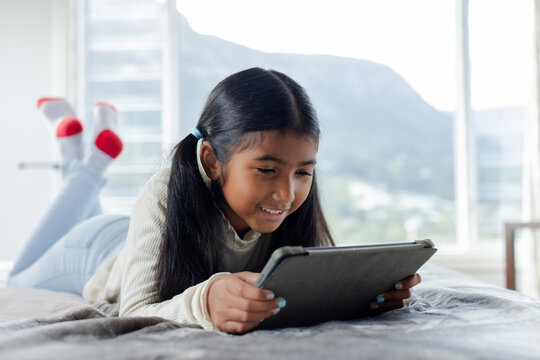 School-age girl using black tablet while lying on bed with gray blanket in bedroom near window
