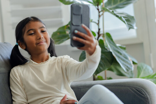 Asian child girl holding smartphone recording selfie on home sofa by shutters and plant, copy space