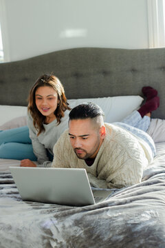 Hispanic couple lying on bed with grey bedspread and white pillows using silver laptop in bedroom
