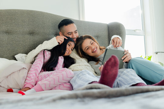 Diverse family of three reclining on bed in pajamas against grey headboard, sharing tablet