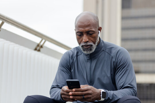 Senior african american man viewing smartphone on rooftop bench by railing using earbuds