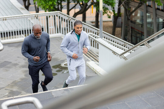 Diverse male partners jogging up wet stairs in running shoes and fitness tracker by metal rails
