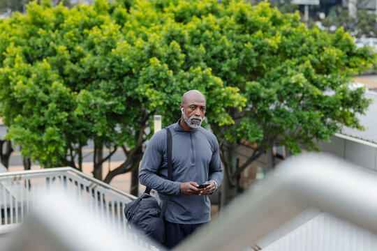 African american man checking phone using earbuds, carrying duffel bag on walkway by metal railings