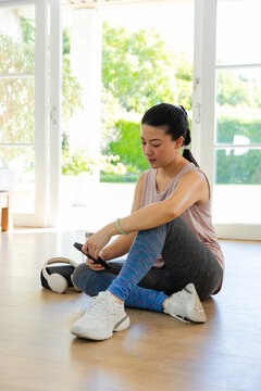 Asian woman sitting on wood floor by doors holding smartphone headphones wearing tanktop leggings