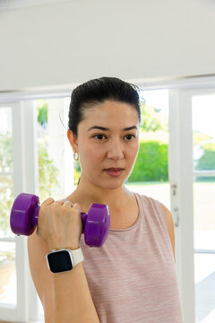 Lifting woman after watch buzzing, purple dumbbell at home by glass doors, wearing pink top, hoops