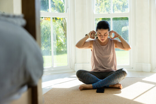 Adult African woman sitting adjusting headphones on woven mat by bay windows, tank, leggings, phone