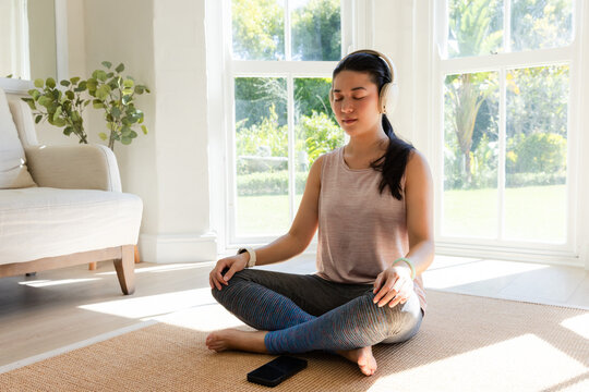 Asian woman meditating cross-legged on rug in living room wearing headphones, smartphone nearby