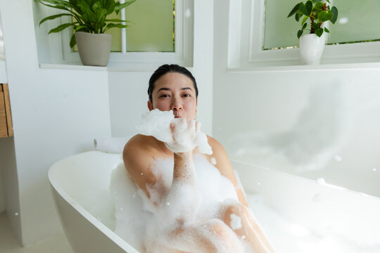 African American woman sitting in freestanding tub, holding foam and blowing bubbles toward lens