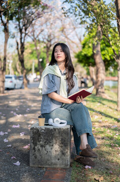 Pretty asian woman looking away from a book while sitting cross legged on concrete bench at sidewalk