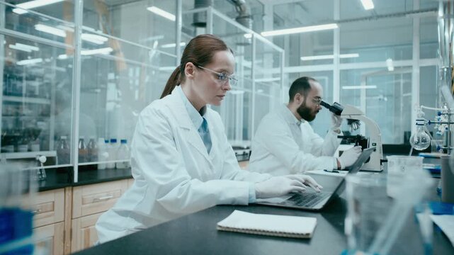 Female scientist in gloves typing on laptop at workplace in modern laboratory as male colleague working with microscope in background