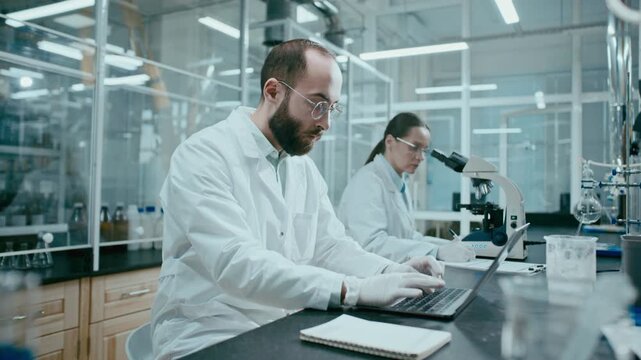 Male scientist in gloves working on laptop at table in modern laboratory, his female colleague conducting microscopic research and taking notes in background