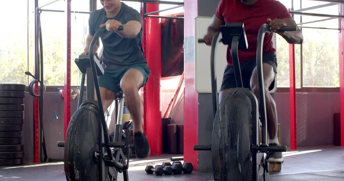 Diverse male workout partners glancing at smartwatch then sprinting on bikes in gym