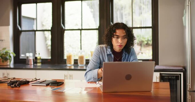 Non-binary person scanning laptop, reaching for pen, jotting notes for work at kitchen
