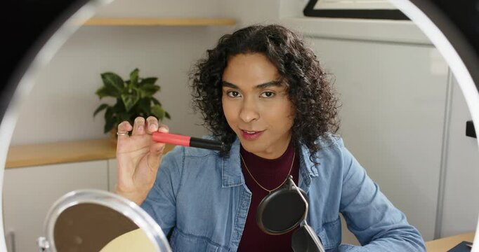 Non-binary creator glancing at mirror showing red lipstick applying lip swatch at desk for review