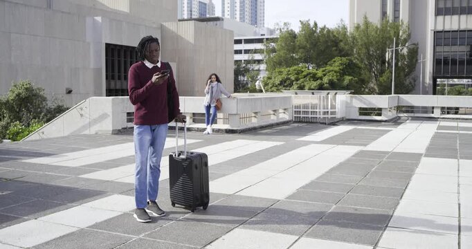 Receiving call, mid adult African American man rolling suitcase on roof terrace glancing at screen