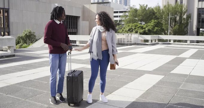 Diverse couple pointing to building, using phone map, planning route and rolling suitcase on plaza