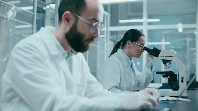 Male scientist typing data on laptop while his female colleague examining specimen under microscope during laboratory research. Rack focus
