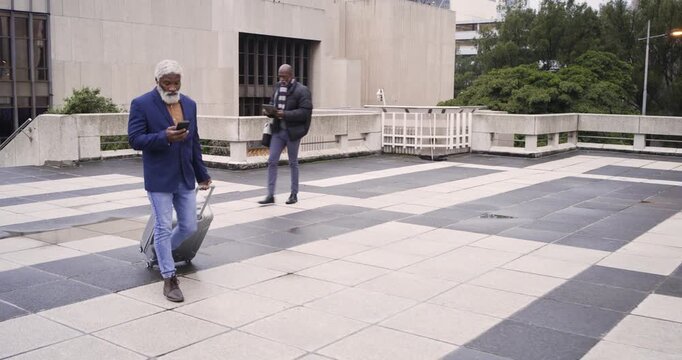 Mature African American man pulling suitcase checking smartphone second man using tablet on plaza