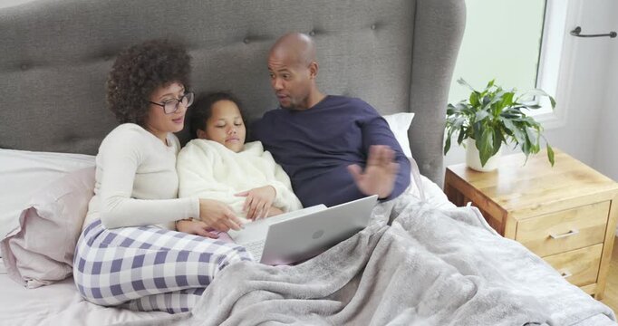 Family settling into bed under gray fleece blanket guiding daughter navigating laptop for learning
