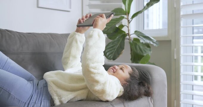 Child girl tapping and swiping tablet on grey sofa under shuttered window, laughing after wobble