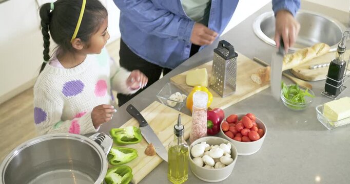 Girl reaching pepper mill in kitchen as adults guiding onion chopping and cheese grating for meal