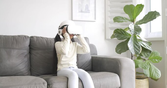 Girl student picking up white VR headset, pulling over eyes, exploring virtual world on grey couch