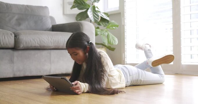 Propping elbows on hardwood floor, Asian child girl tapping tablet while playing by sofa and plant