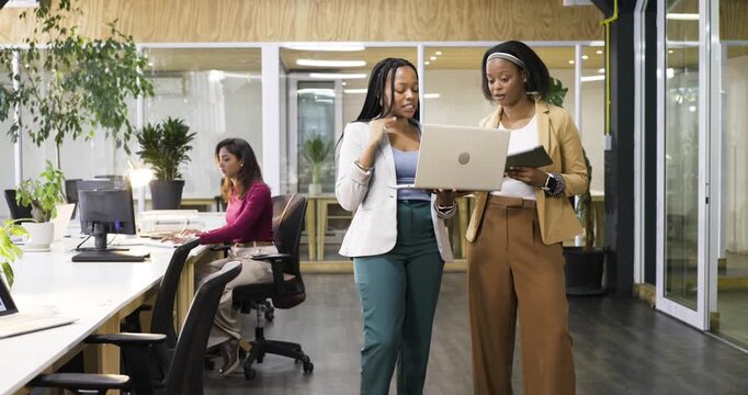 Two African American women walking to office desk, sharing project chart on laptop, tapping tablet