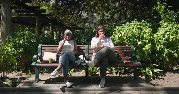 Diverse female friends resting on park bench eating food sipping drinks glancing as people walk