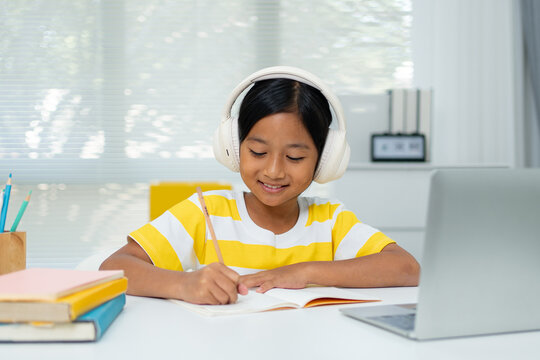 Asian Girl with Headphones Taking Notes While Learning Online on Laptop at Home