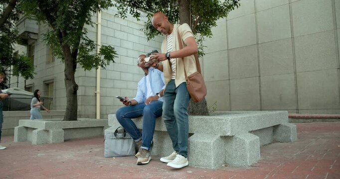 Two African American men greeting, second man approaching showing phone and offering snack at plaza
