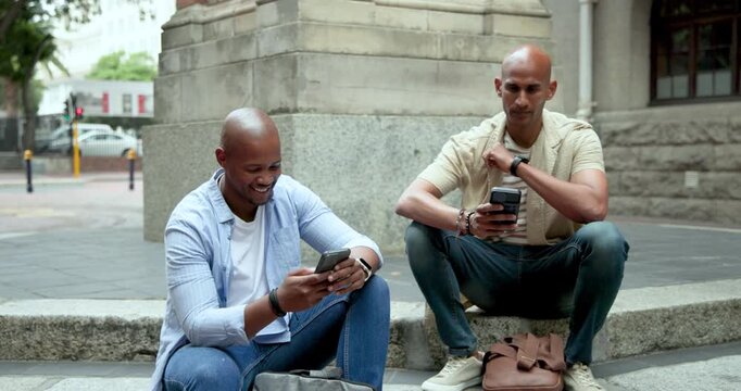 Diverse male friends sitting on ledge at sidewalk, laughing at phone alert and showing screen