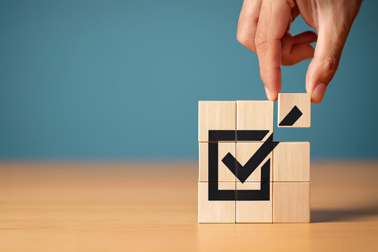 Completion and approval concept. Abstract background with hand placing final wooden block into cube puzzle forming black check mark inside square symbolizing success, validation, and task achievement.