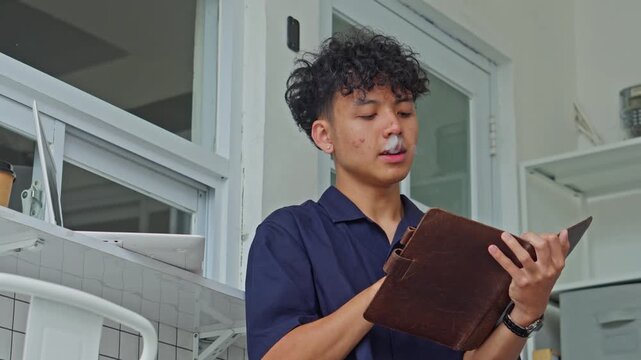 Young man in polo shirt reads from leather journal while smoking cigarette in outdoor setting