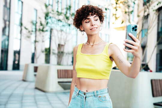 Young beautiful smiling hipster woman in trendy summer clothes. Carefree woman with curls hairstyle, in the street. Positive model holds mobile phone, looks at cellphone screen, uses smartphone apps