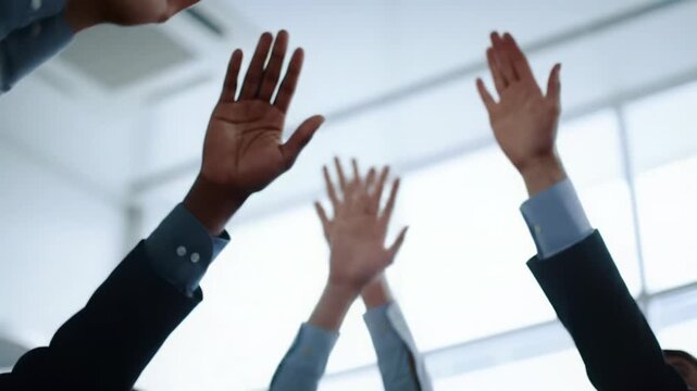 Low-angle video of diverse human hands interlocking in a brightly lit modern office, smoothly dollying in then tilting upward towards light with bokeh background. Concept of upward progress