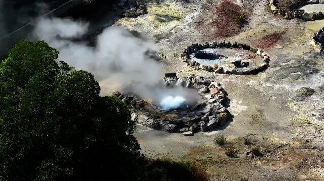 Aerial view of Furnas fumaroles where steam rises from geothermal vents, surrounded by dark rocks and contrasting volcanic textures in the Azores, Portugal.