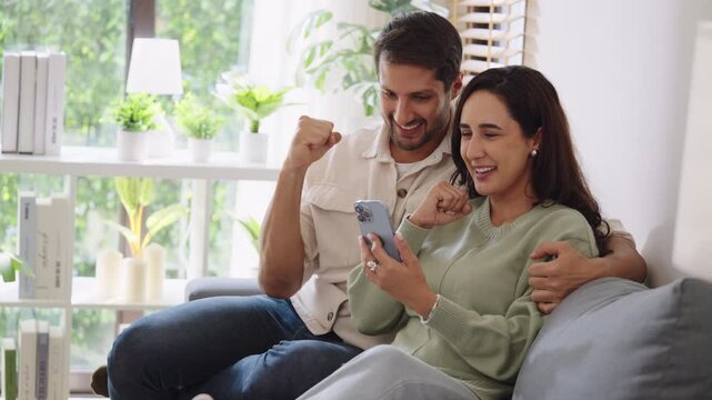 Hispanic couple using mobile device watching social media and winnning prize online at home on the sofa
