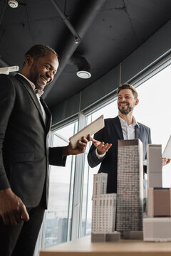 Two smiling businessmen in suits review architectural models of skyscrapers in a modern office with a large window overlooking a city skyline