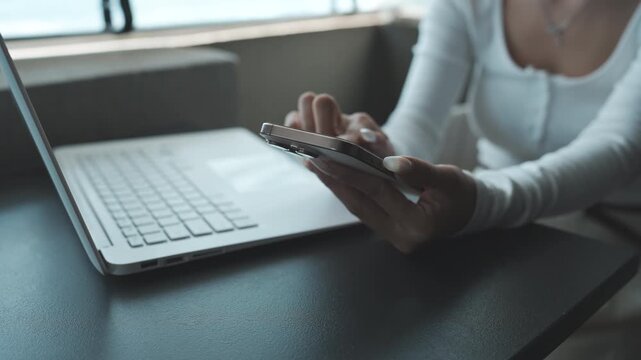 Close-up of a person using a smartphone beside a laptop on a desk, natural light highlighting a modern workspace and multitasking digital lifestyle.
