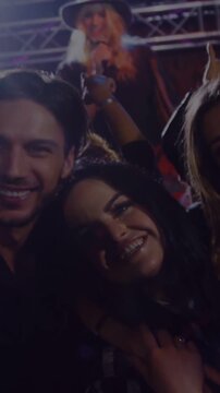 Vertical video: Duo leaning at concert, friend raising phone capturing selfie with stage lights