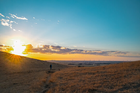 Landcape view of a woman hiking along a dirt trail in a dry, grassy field during a scenic sunset with distant wind turbines on the horizon, Mount Noorat, Victoria, Australia
