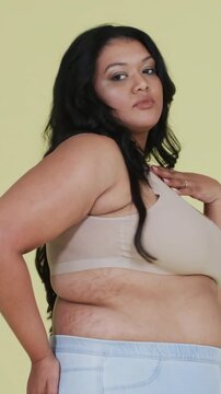 Vertical video: Posing woman from left, fixing hair, turning in studio, showing bralette and denim