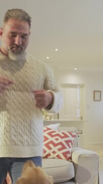 Vertical video: Gesturing man in cream knit sweater offering treat near couch, making dog jump