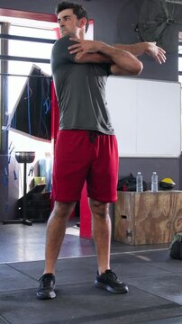 Vertical video: Starting warm-up man in grey tee red shorts doing arm stretches at gym with bottles