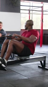 Vertical video: Rowing two men red shirt leading, starting at catch pulling on ergs at gym training