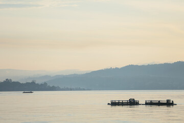 Traditional fishing boat and floating fish farm cages on a calm sea near misty coastal hills © anwar