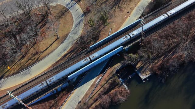 An aerial view of a Long Island Rail Road train traveling on a sunny day. The drone camera tilted downward is stationary but zooms in slowly, as the train passes by dry leafless trees.
