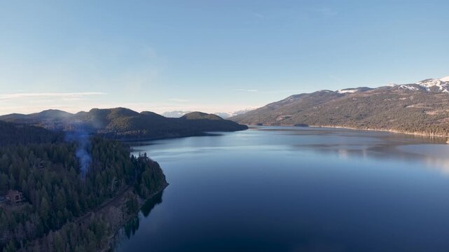 Aerial view of a calm blue lake bordered by dense evergreen forests with a plume of smoke rising near the shore and rolling hills and farmland under a clear sky.