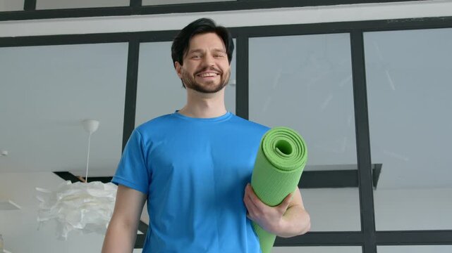 Portrait of active motivated handsome sporty fit man holding rolled up exercise mat, looking with cheerful smile, expressing confidence and positivity at home.